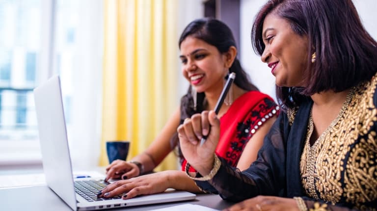 Image of two indian women in Sari's sat at a desk, smiling whilst using a laptop